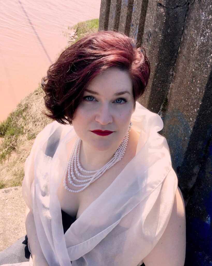 Erin Bardua, a white woman with wavy dark hair, wearing a black dress and white wrap with multiple strands of white pearls. She's sitting on a broken concrete bridge with a muddy river in the background.