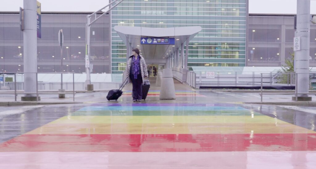 Wide shot of Erin with luggage in a winter coat, crossing a roadway at an airport with a rainbow painted crosswalk. This is a still from the opera film "December"
