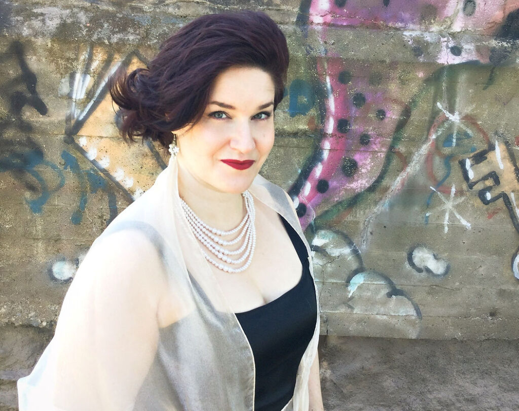 Photo of Erin in front of a graffiti-covered concrete wall. She's a white woman with bobbed dark hair, red lipstick, and a black sleeveless dress with a white wrap. 