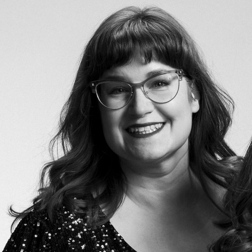 Black and white photo of Erin, a while woman with long wavy hair and fringe wearing glasses and a sparkly dark top. She's smiling energetically at the camera during a fun impromptu photo shoot. 