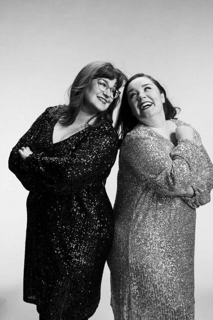 Erin Bardua and Maureen Batt in similar sequined dresses, arms folded and laughing like they're on the poster for a buddy comedy. B&W.